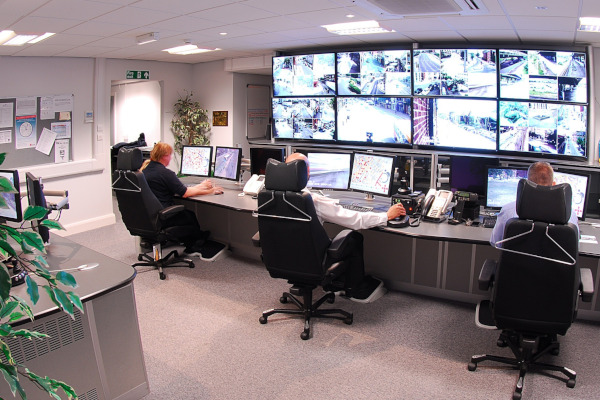 3 Personen auf Drehstühlen die vor einer Wand mit Videos in einem Überwachungsraum sitzen